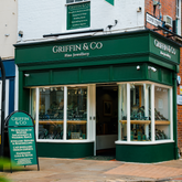 A street-level view of the Griffin & Co Fine Jewellery shop front. The elegant dark green facade features large display windows and its name in silver lettering. An A-frame sign on the pavement lists their specialities.